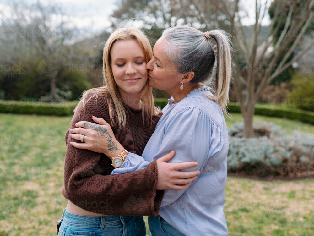 Mum hugging teenage daughter giving her a kiss on the cheek in the backyard - Australian Stock Image