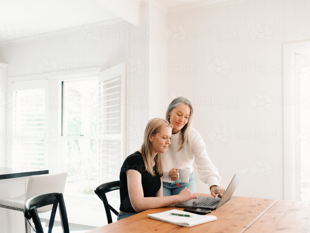 Mum leaning over teenage daughter working on her laptop on the wooden table - Australian Stock Image