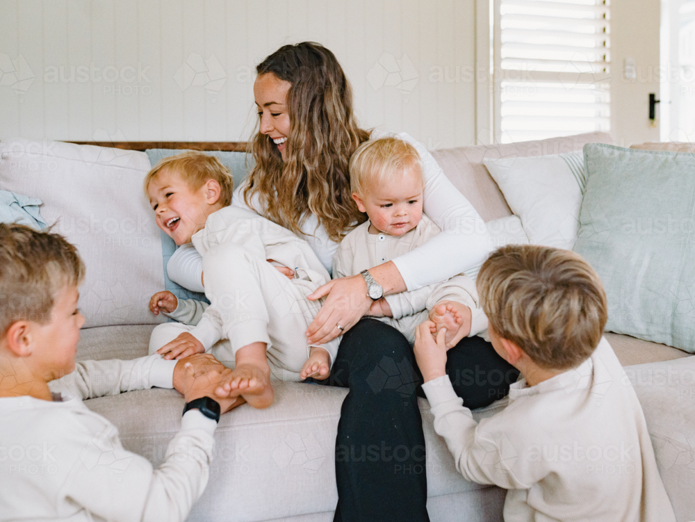 Mum sitting on the couch cradling her two sons while older brothers tickling their feet - Australian Stock Image