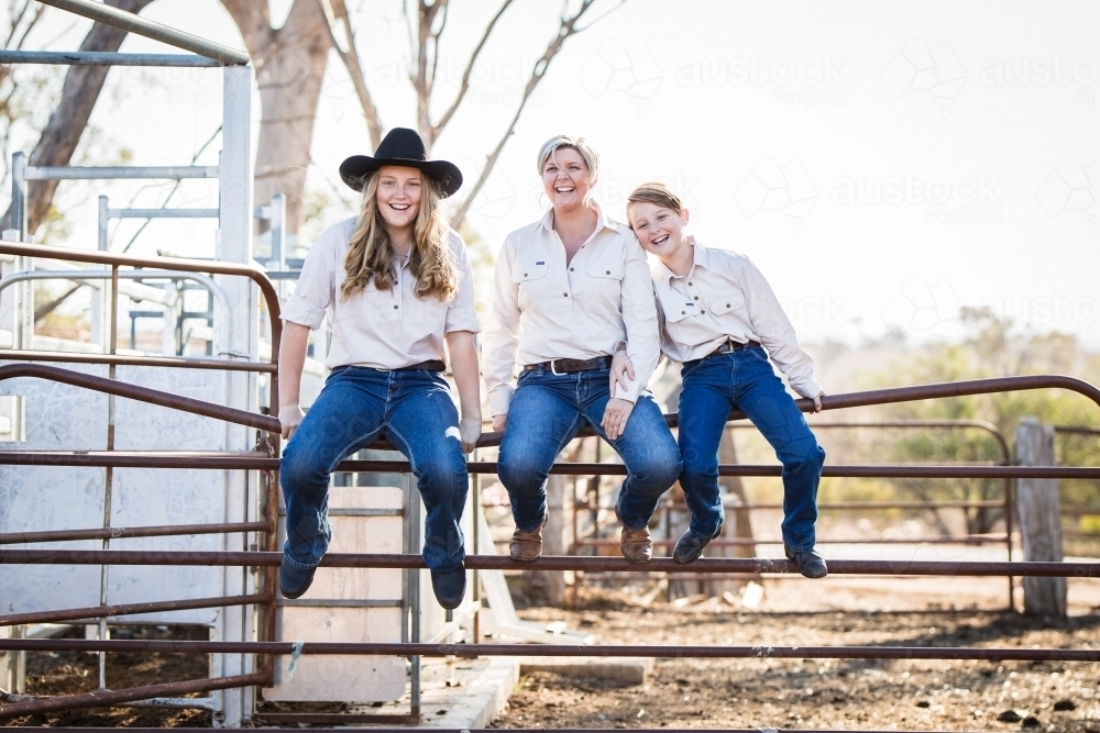 Mum sitting with children on fence in yards on farm in drought - Australian Stock Image