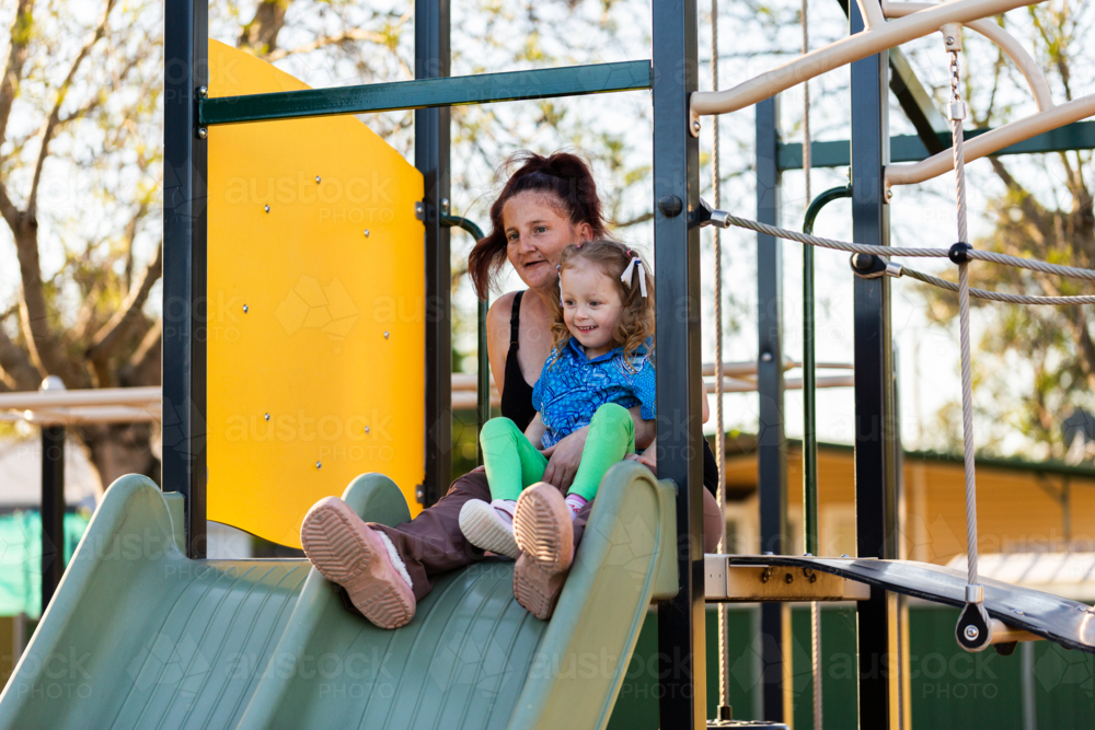 Mum with daughter at playground sliding down slide together - Australian Stock Image