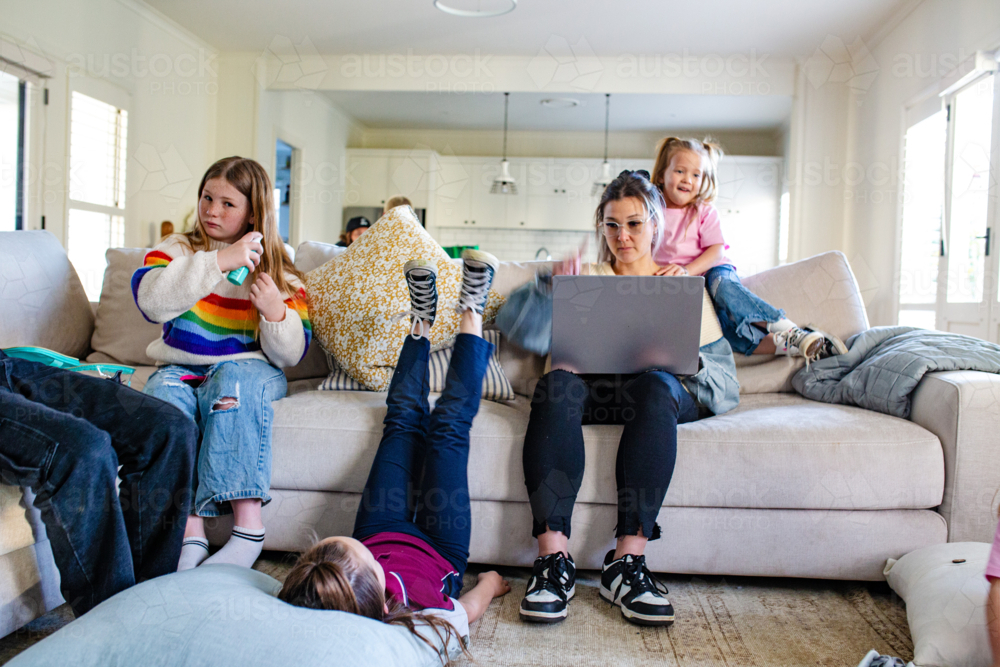 Mum working on her laptop on the couch with daughters playing around the living room - Australian Stock Image