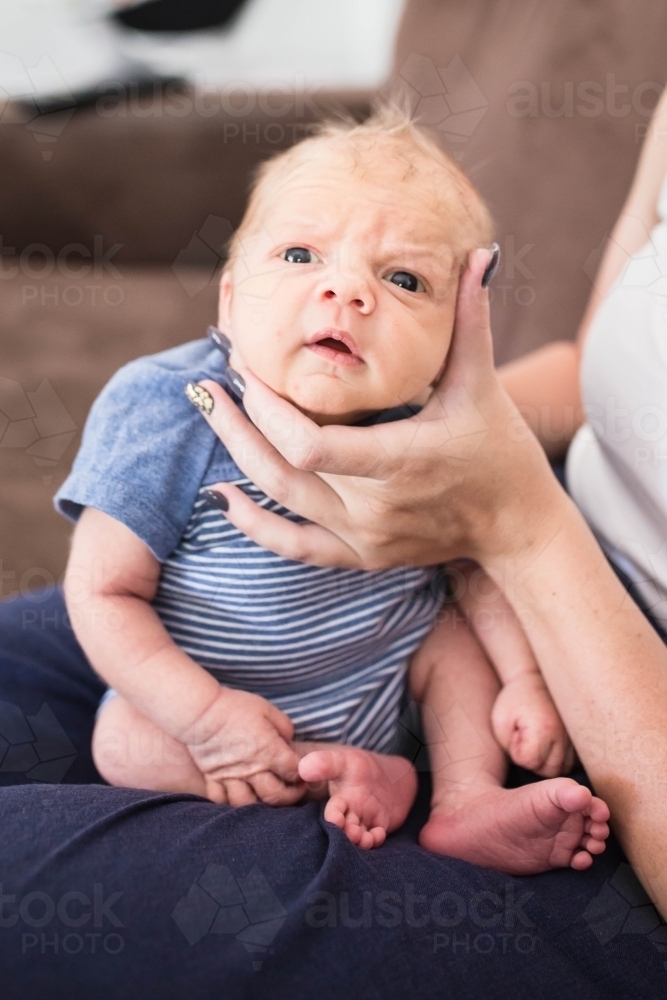 Newborn baby boy being burped on mother's lap while mum holds his face - Australian Stock Image