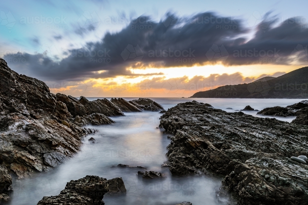 Ocean and rocky coastline against cloudy sunrise - Australian Stock Image