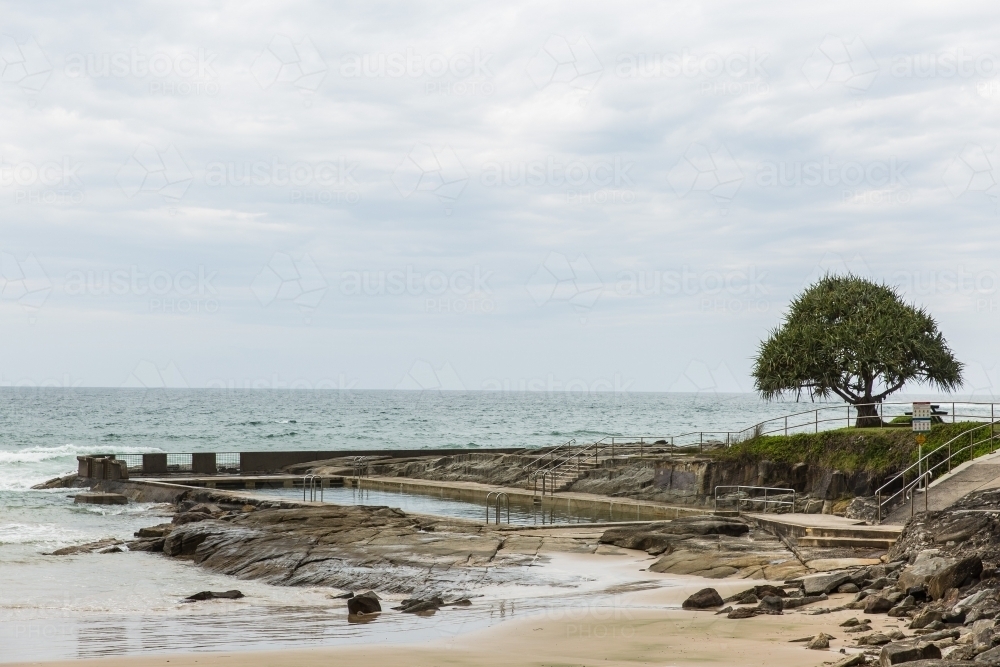 Ocean pool on beach with rocks and sand and tree with cloudy skies - Australian Stock Image