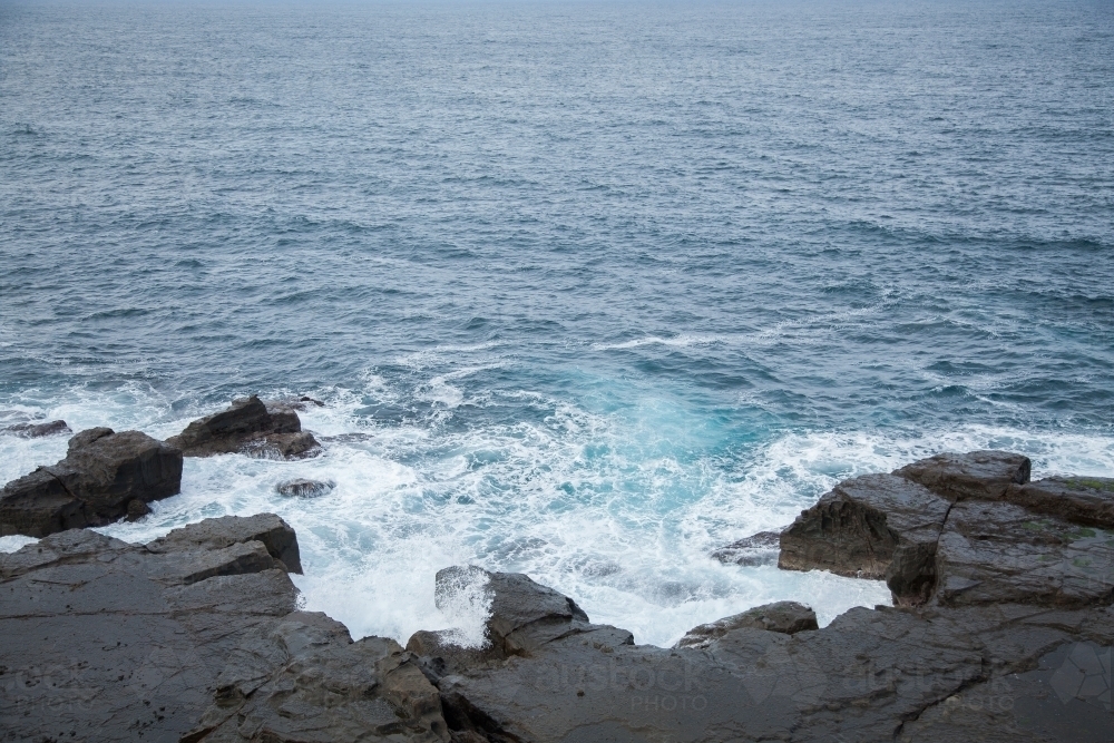 Ocean pounding against black rocks at the base of a cliff - Australian Stock Image
