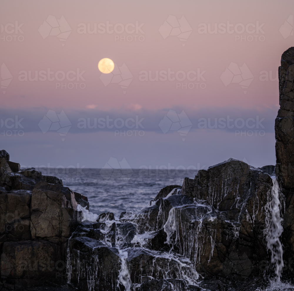 ocean water spilling down rocks with full moon rising in background - Australian Stock Image