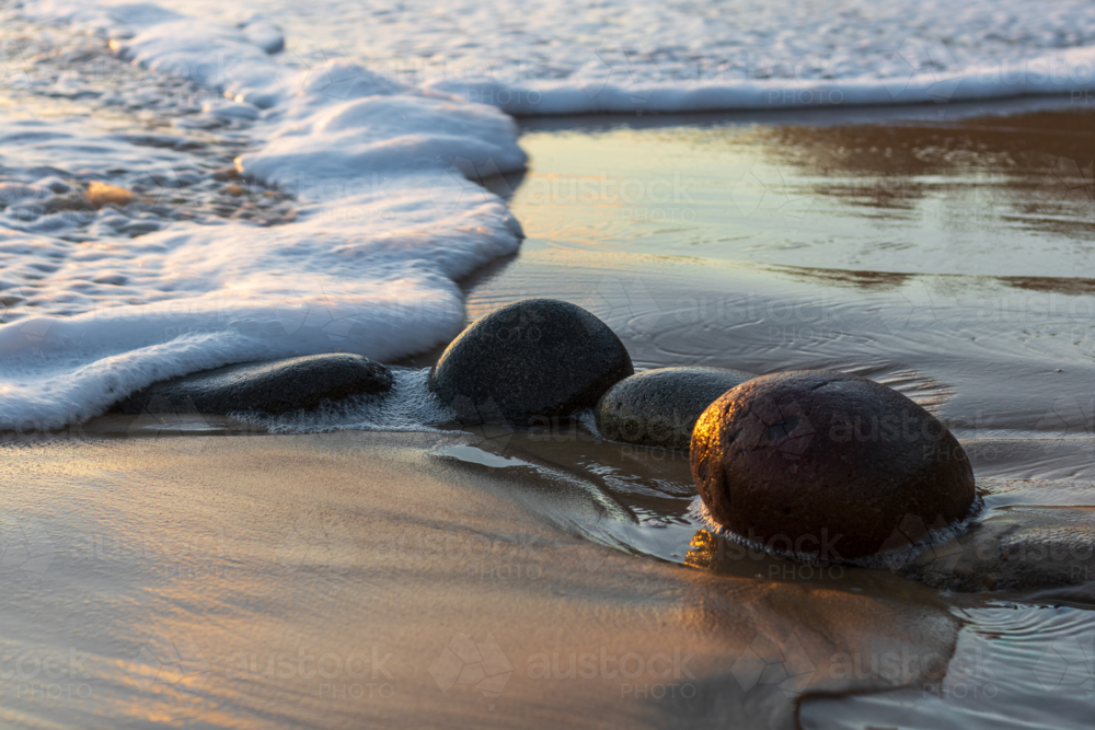 Ocean wave with sea foam washing over smooth round rocks on wet sand - Australian Stock Image