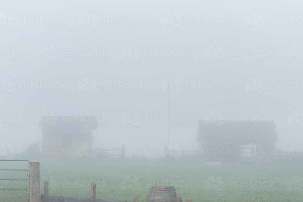 Old farm buildings barely visible through fog on misty morning - Australian Stock Image