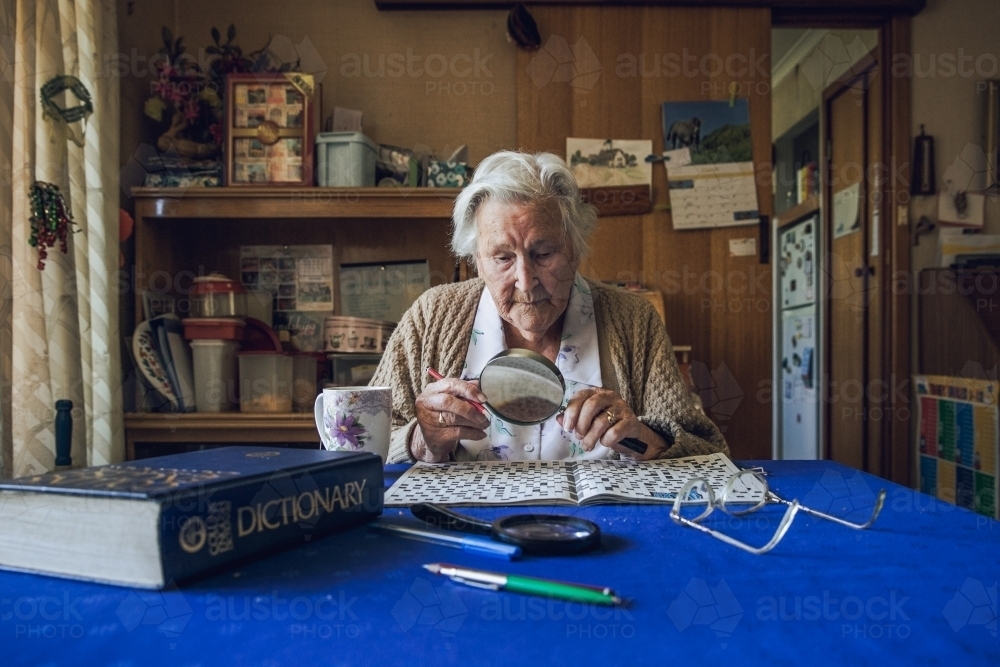 Old lady at dining table doing crossword puzzle with dictionary, magnifying glass, glasses - Australian Stock Image