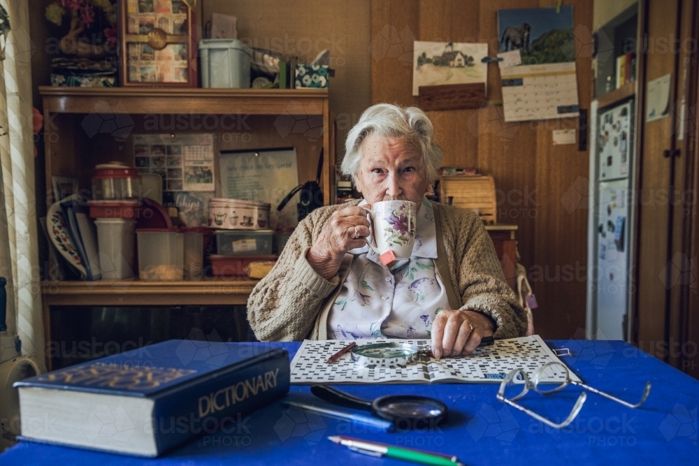 Old lady at dining table drinking tea with crossword, magnifying glass, dictionary - Australian Stock Image