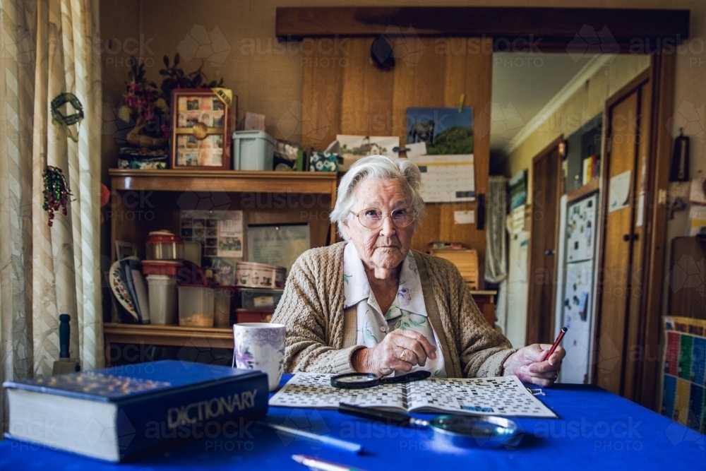 Old lady at dining table with crossword, magnifying glass, dictionary and cup of tea - Australian Stock Image