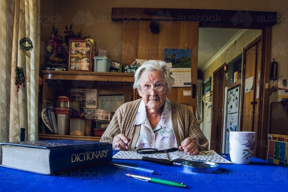 Old lady at dining table with crossword, magnifying glass, dictionary and cup of tea - Australian Stock Image