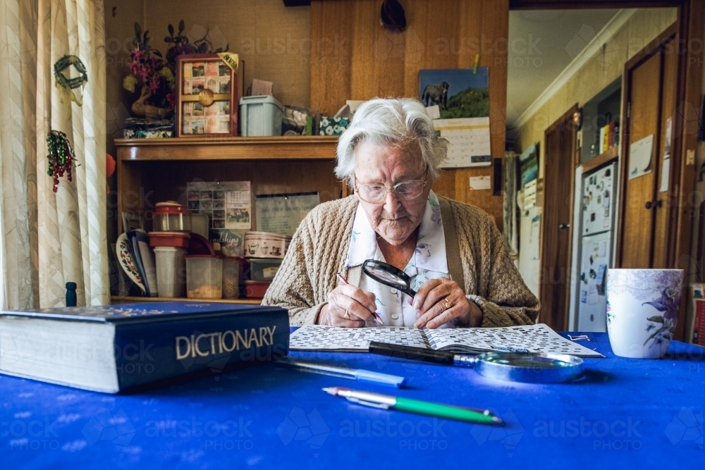 Old lady at dining table with crossword, magnifying glass, dictionary and cup of tea - Australian Stock Image
