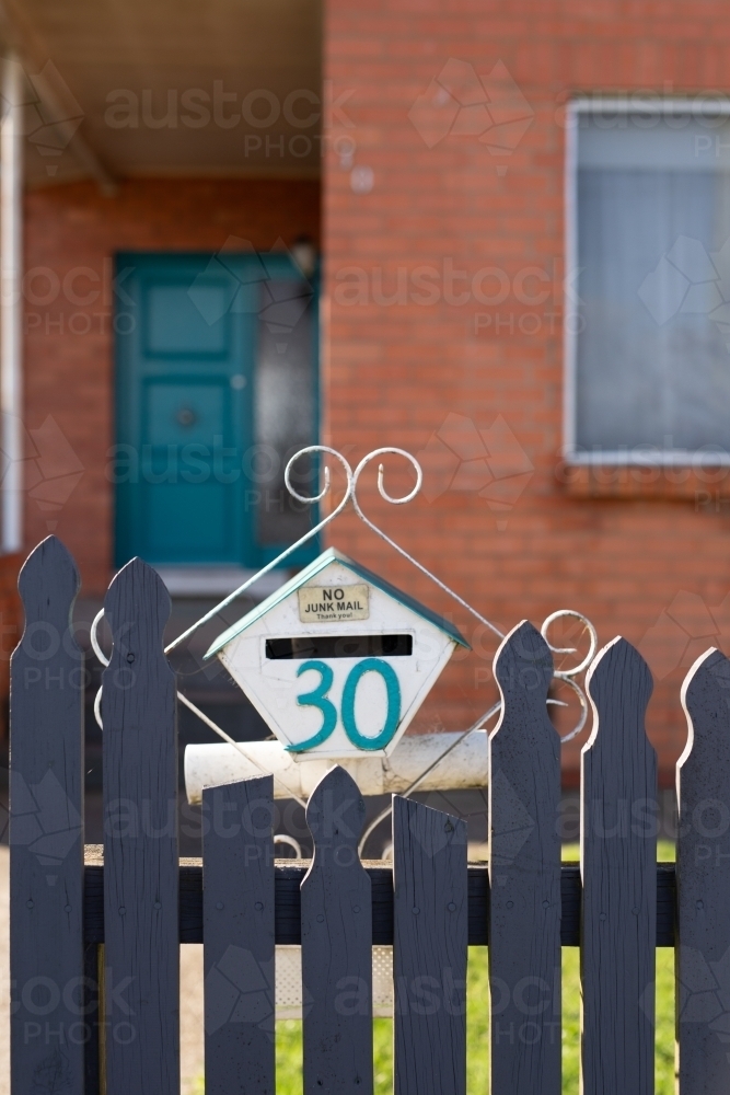 Old letterbox on blue picket fence with red brick house behind - Australian Stock Image