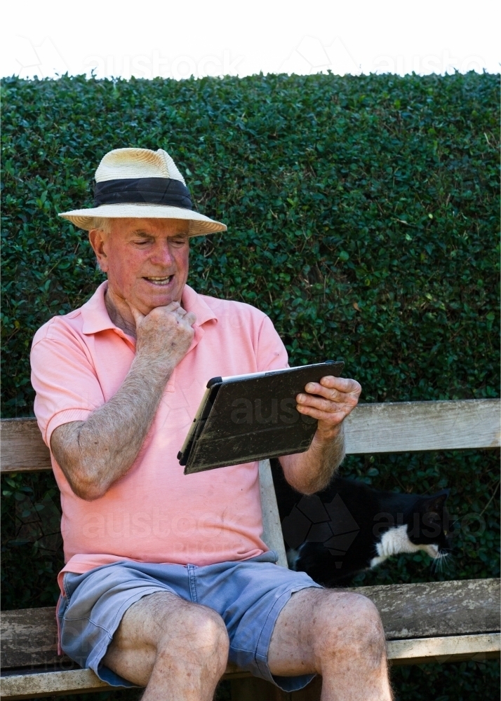 Old man sitting on a bench looking at his tablet - Australian Stock Image
