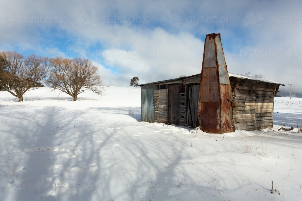 Old timber shack, shed or stable in a rural paddock with full coverage snowfall in winter. Blue sky - Australian Stock Image