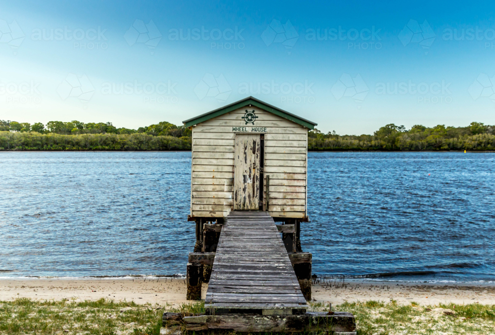 Old weathered wooden boat house with crooked jetty on river with blue sky and green vegetation - Australian Stock Image