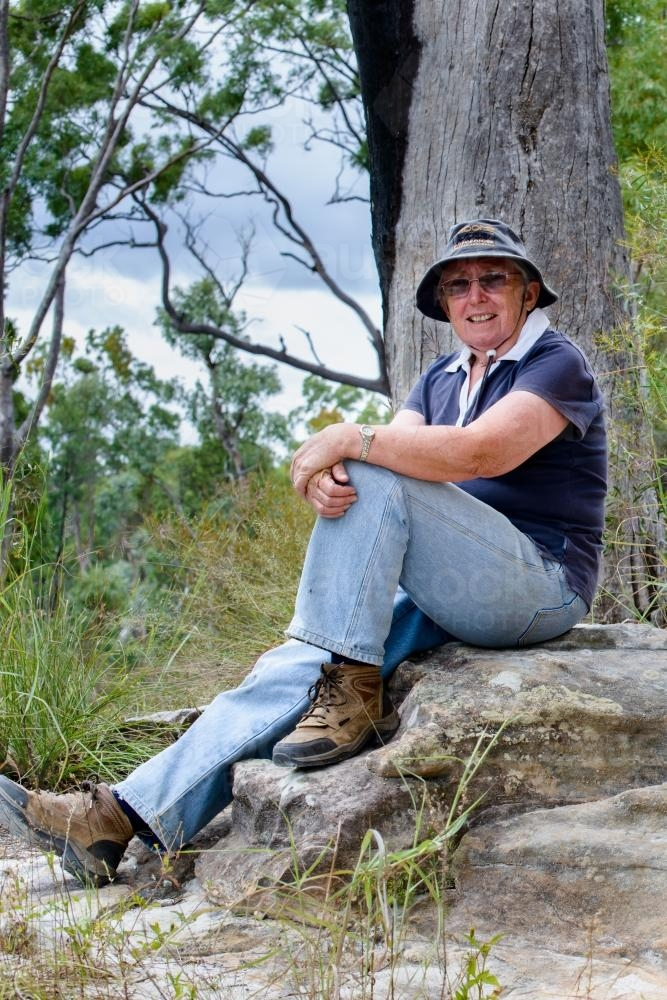 Old woman sitting on rock in remote, rugged setting - Australian Stock Image