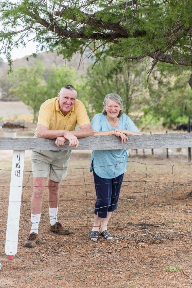 Older couple husband and wife retirees leaning on fence post at home on farm - Australian Stock Image
