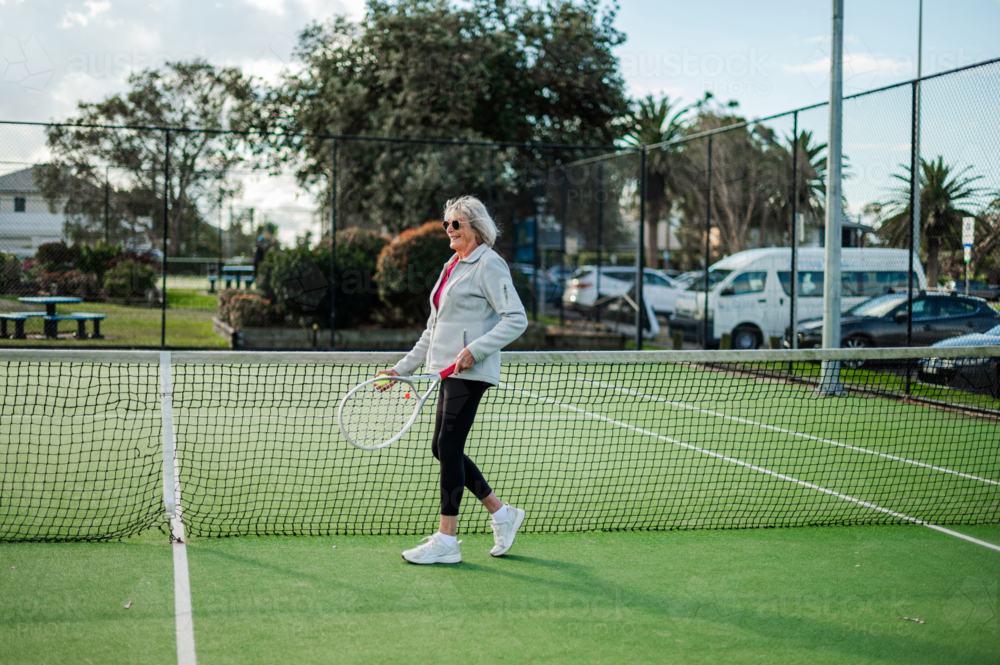 Older female athlete walks on a tennis court, enjoying an afternoon match. - Australian Stock Image