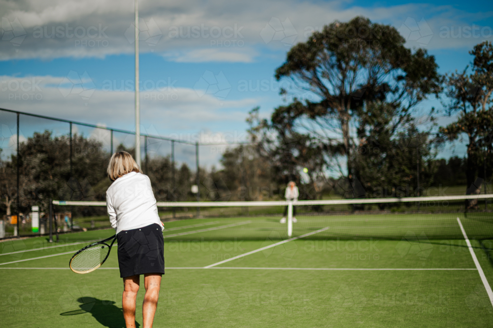 Older female tennis player serves on a maintained green outdoor court - Australian Stock Image