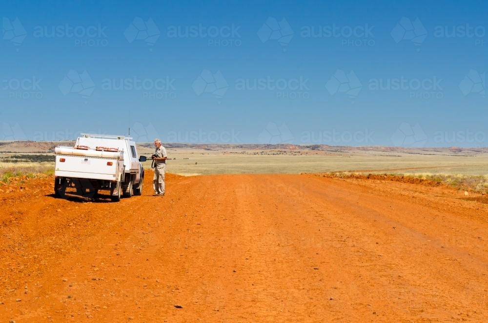 Older grey haired man beside vehicle towing a trailler on side of remote wide orange dirt road - Australian Stock Image