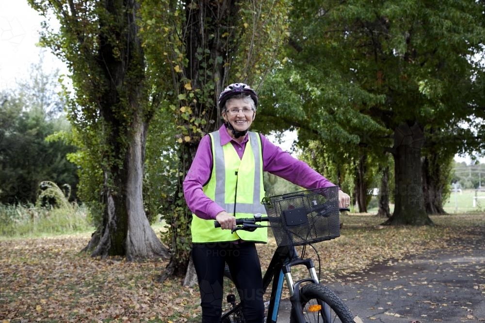 Older lady wearing high visibility vest and helmet standing with bike - Australian Stock Image