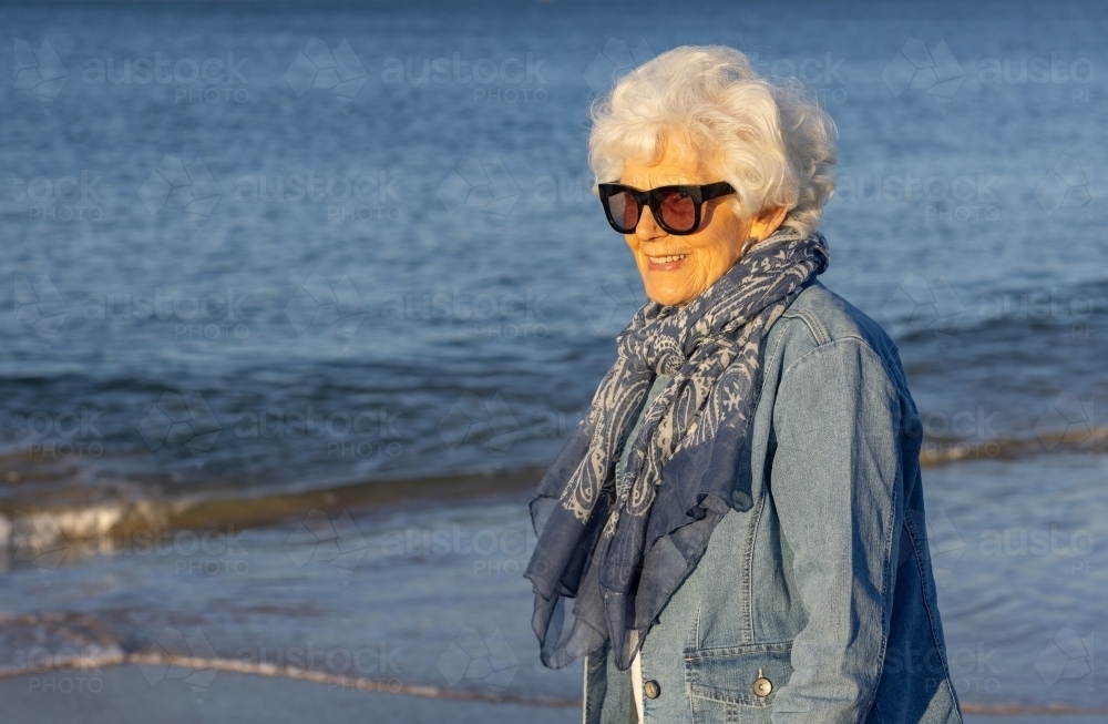 older lady wearing sunglass with scarf and denim jacket on the beach - Australian Stock Image