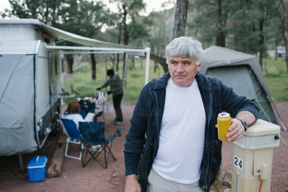 Older man standing in front of a caravan and tent with a beer - Australian Stock Image