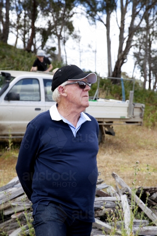 Older man standing in front of wood pile on a farm looking away into the distance - Australian Stock Image