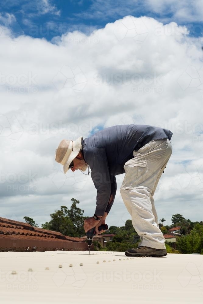 Older man with grey beard working with drill on house roof - Australian Stock Image