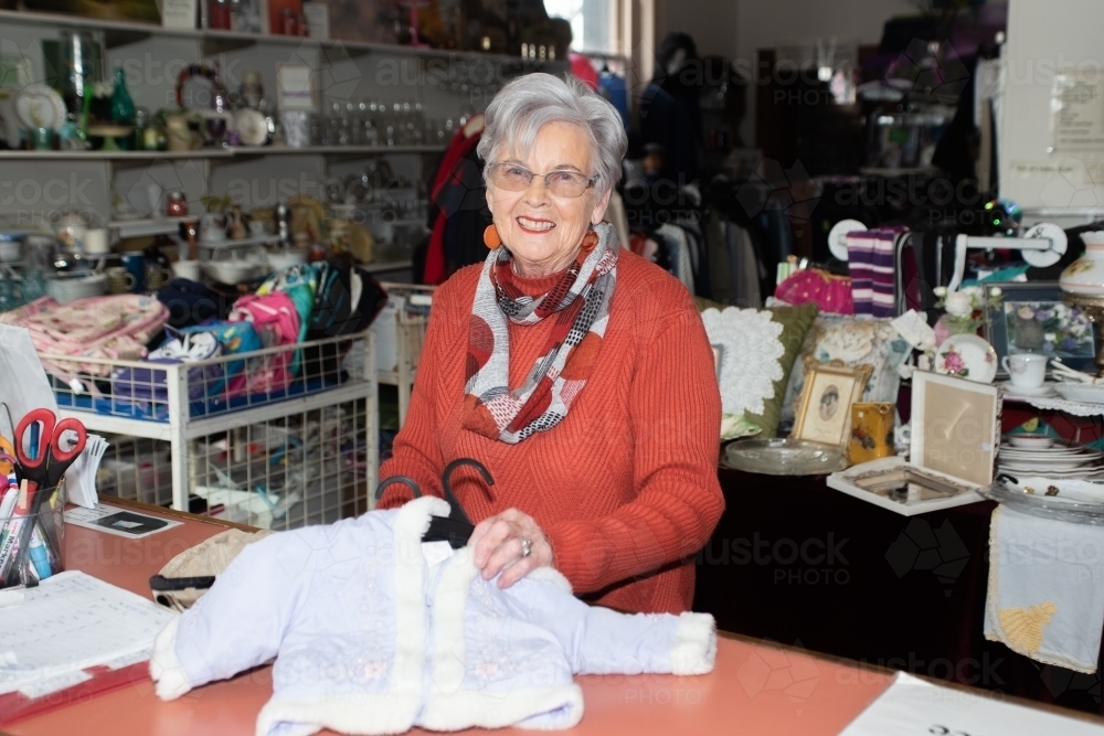 Older smiling lady showing baby clothes in an op shop - Australian Stock Image
