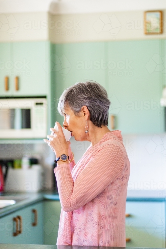 Older woman drinking a warm tea beverage in pastel kitchen - Australian Stock Image