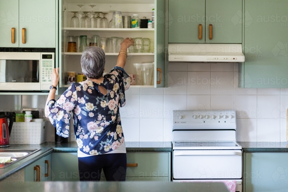 Older woman reaching for water glass in kitchen cupboard - Australian Stock Image
