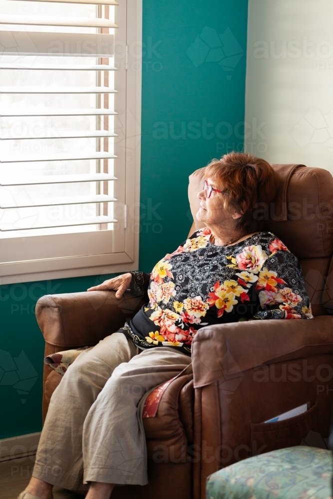 older woman sitting in chair looking dreamily out window - Australian Stock Image