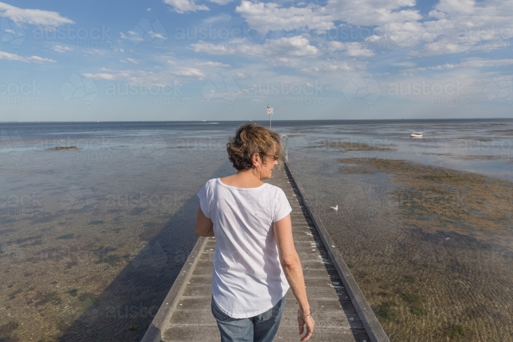 Older woman walking on timber pier looking over tidal shallows - Australian Stock Image