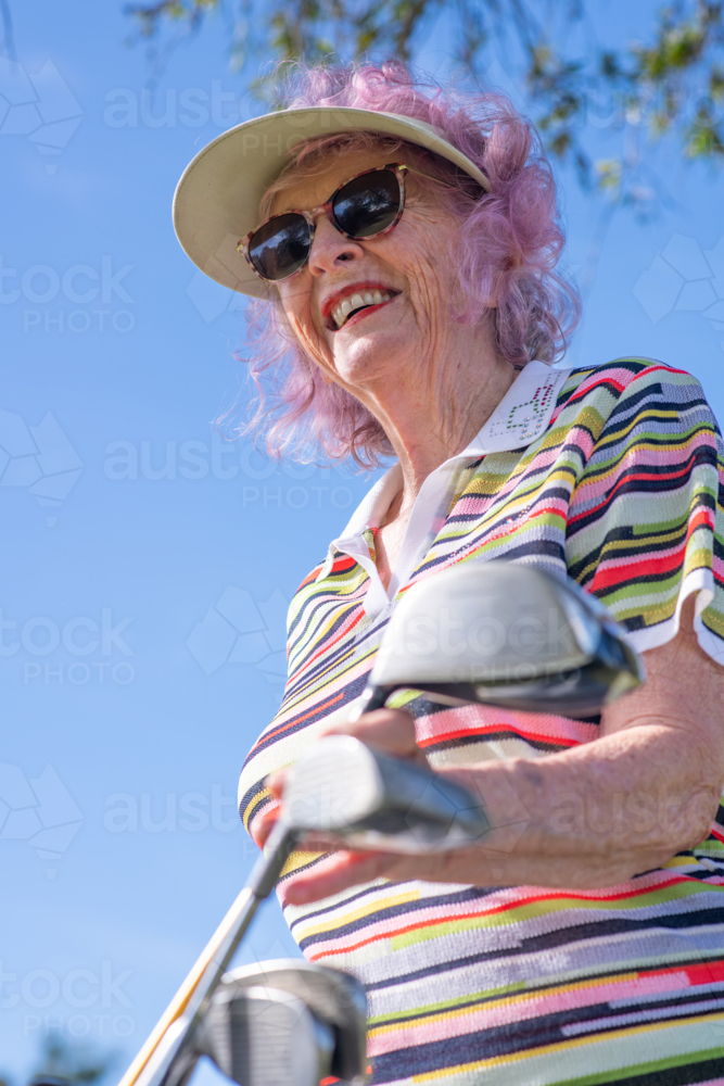 Older woman with pink hair wearing visor and striped shirt standing near golf clubs outdoors - Australian Stock Image
