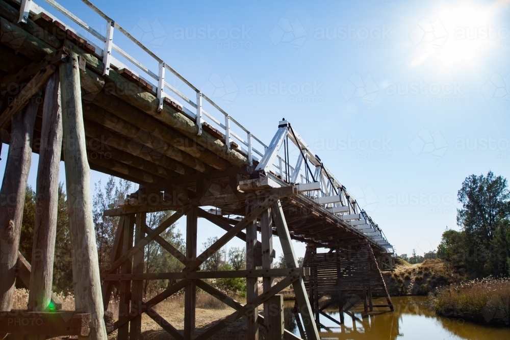 Sun flare over pillars of timber truss road bridge above creek - Australian Stock Image