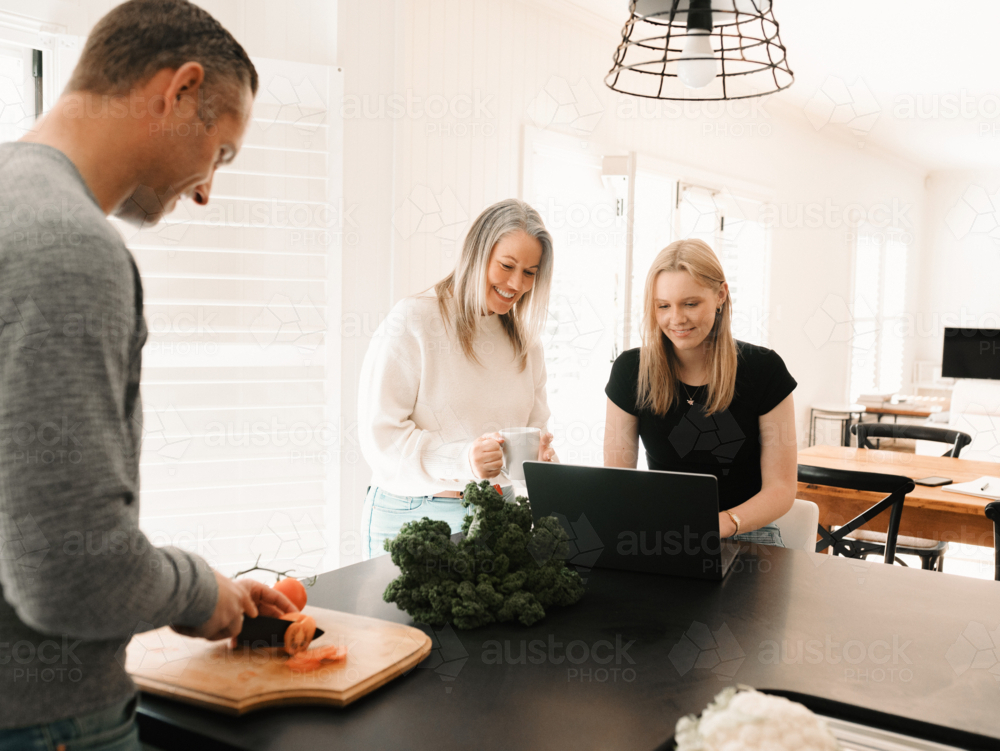 One male adult preparing food while mum helps teen daughter with work on laptop in bright kitchen - Australian Stock Image