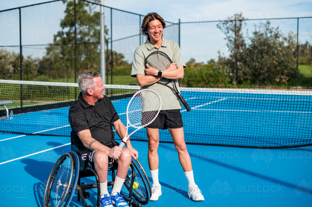 One player sits in a wheelchair while the other stands, both smiling and holding racquets - Australian Stock Image