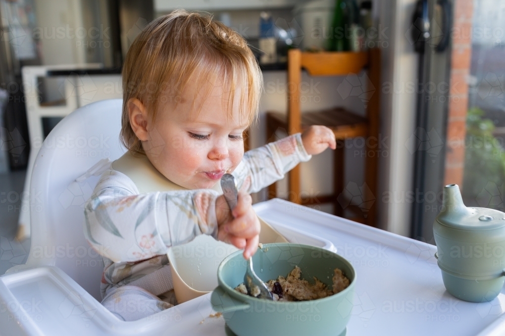 Image of One year old baby girl using spoon to eat weetbix and sultanas for breakfast Austockphoto