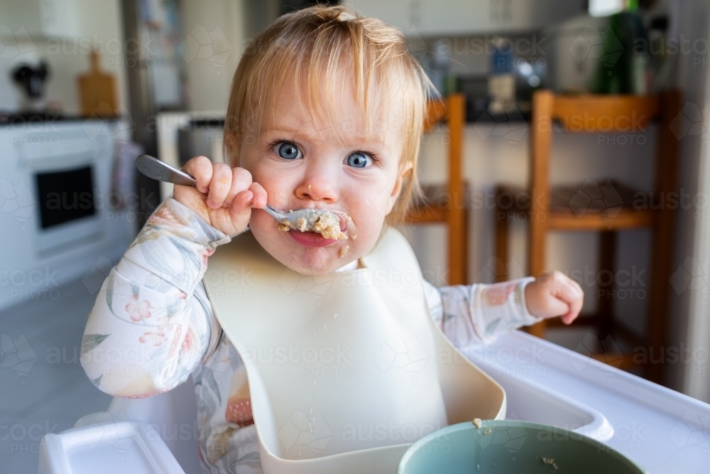 Image Of One Year Old Baby Girl Using Spoon To Eat Weetbix And Sultanas image-of-one-year-old-baby-girl-using-spoon-to-eat-weetbix-and-sultanas