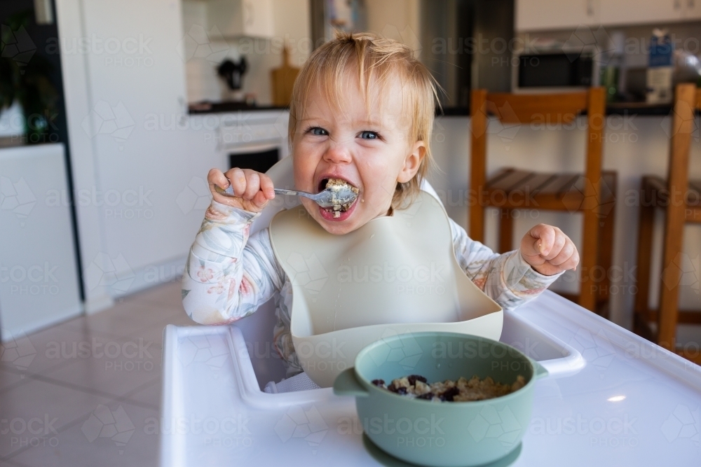 Image of One year old baby girl using spoon to eat weetbix and sultanas for breakfast Austockphoto