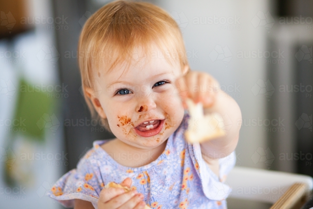 One year old baby with messy face holding out piece of toast to share - Australian Stock Image