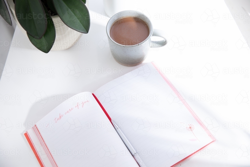 Open journal on a white desk with a hot beverage in a mug in morning light - Australian Stock Image