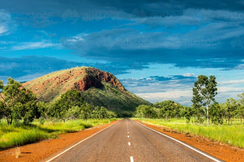 Open road leading to a red rocky hill under a dramatic sky with eagle flying overhead - Australian Stock Image