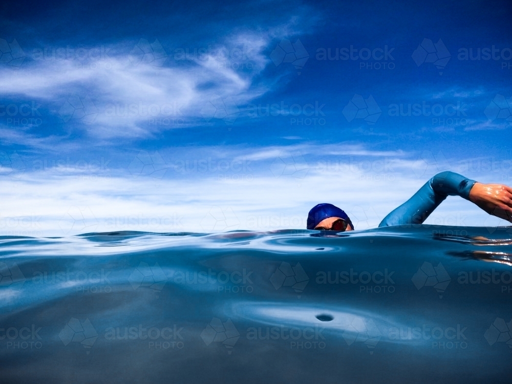 Open Water female swimmer doing freestyle  in glassy ocean wearing goggles, wetsuit and cap - Australian Stock Image