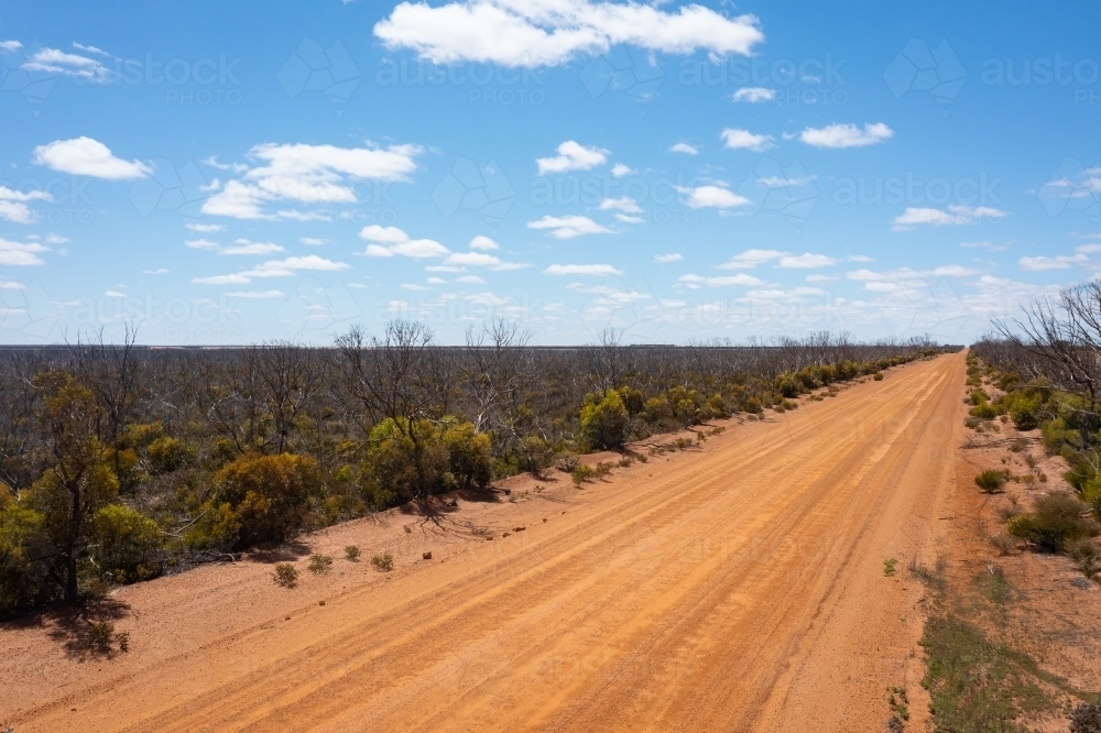 orange gravel road stretching to horizon with low scrub and blue sky - Australian Stock Image