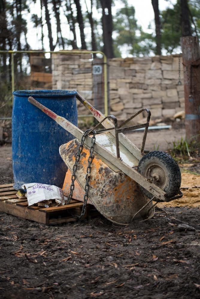 Orange wheelbarrow and blue barrel on pallet in front of drystone wall - Australian Stock Image